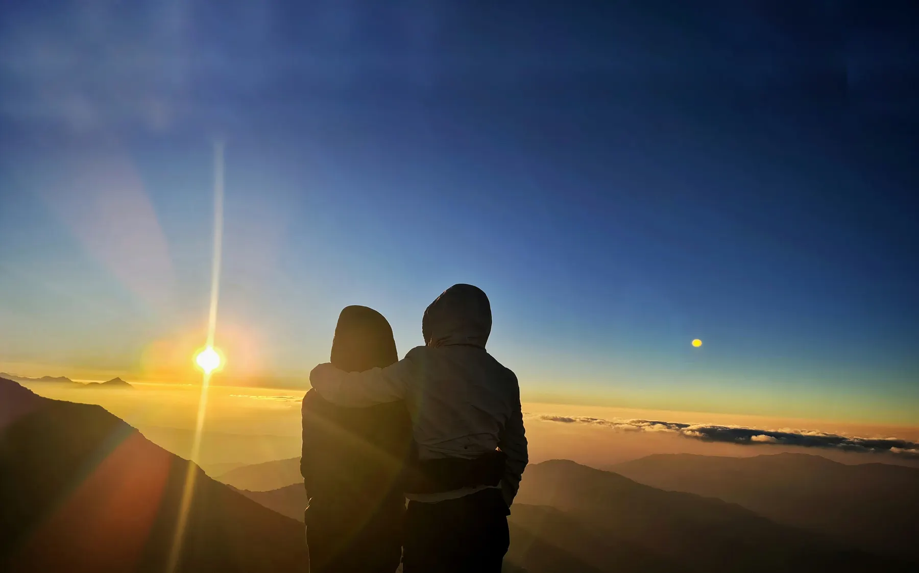 Two people silhouetted against a wide golden sunrise over distant hills