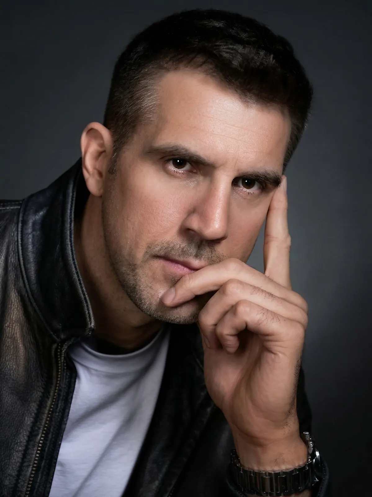 Man in a leather jacket resting his chin on his hand against a dark background