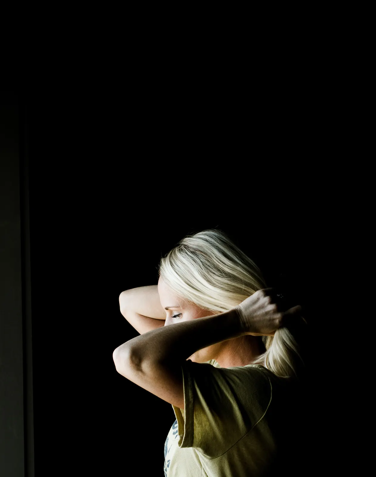 Studio portrait of a woman tying her hair back, turned away from the camera against a dark background