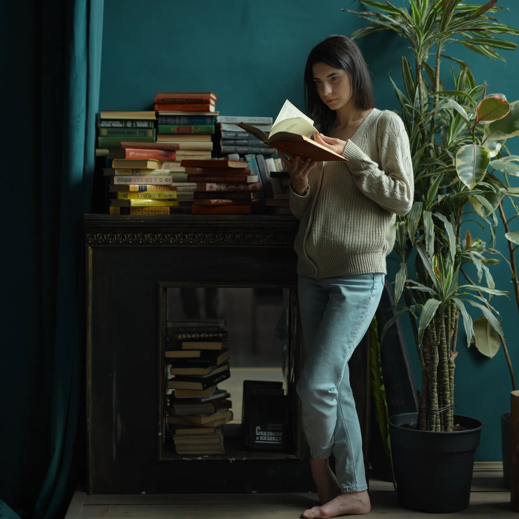 Woman reading beside a bookshelf and houseplants in a dark green room